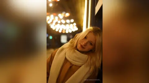 Blonde woman in a cozy brown coat and white scarf smiling under warm city lights at night.
