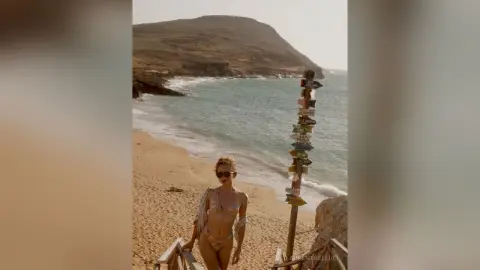 LaurentBelluci stands on a sandy beach wearing a light-colored bikini and sunglasses with a wooden signpost behind her.