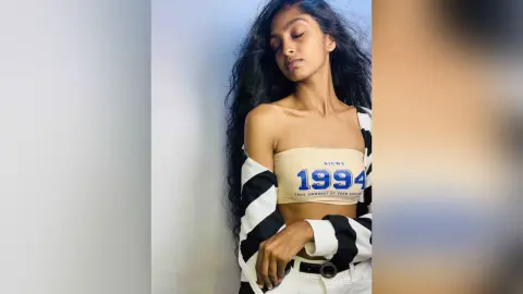 Young woman with long wavy hair wearing a beige crop top and striped jacket posing against a light wall.