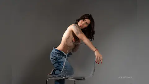 AlesiaNoir — Tattooed woman in denim jeans sitting on a clear chair against a grey studio background in a moody pose.