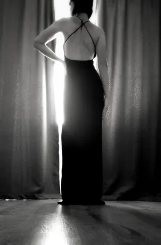 Elegant woman in a backless black evening gown standing by backlit curtains in a moody black and white setting.