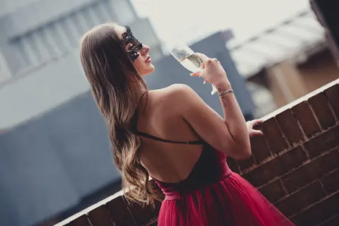Elegant woman in a red evening dress and black masquerade mask sipping champagne on a rooftop at night.