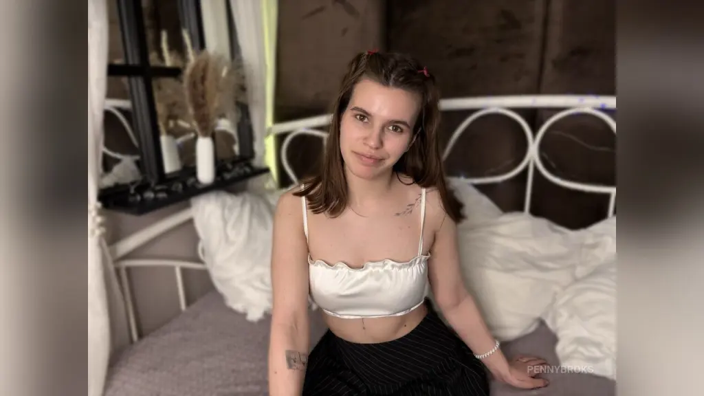 Young woman with chest tattoos posing by a bright window in a natural, moody indoor setting.