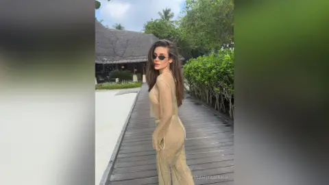 Stylish woman in a gold mesh cover-up walking on a wooden boardwalk at a tropical beach resort.
