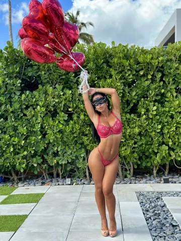 Woman in red lace bikini holding pink balloons in a lush tropical garden setting.