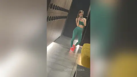 Fit woman in green gym set taking a mirror selfie in a modern locker room setting.