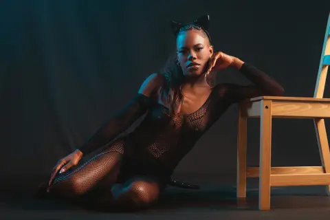Moody portrait of a woman in a black mesh bodysuit and cat ears sitting on the floor by a wooden chair.
