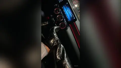 Hailey wears shiny black leather boots while sitting in a car with red-lit dashboard gauges.