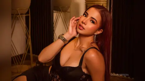 Glamorous woman in a black lace outfit posing with a confident expression in warm, moody indoor lighting.