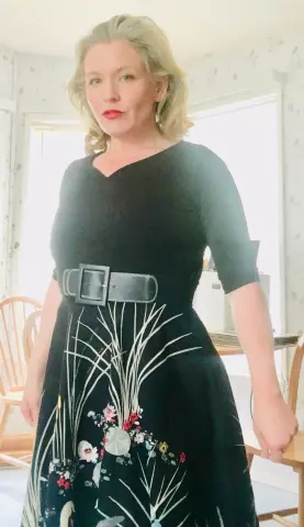Blonde woman in a black floral midi dress with a wide belt standing in a bright, sunlit room.