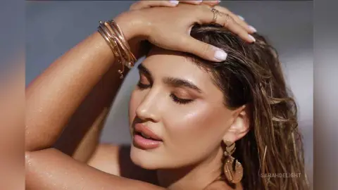 Close-up of a woman with wet hair and gold jewelry posing in warm, glowing sunlight.