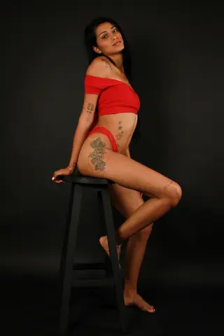 Tattooed woman in red crop top and bikini posing on a stool against a black studio background.