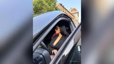 NicoleBella sits in a car wearing a black blazer and sunglasses, looking off to the side.
