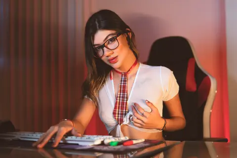 Young woman with glasses in a white crop top sitting at a desk in a moody, dimly lit room.