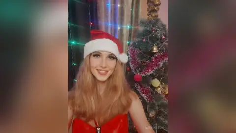Smiling blonde woman in a red Santa hat and festive outfit posing by a decorated Christmas tree.