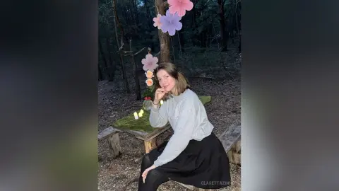 Young woman in a grey sweater and black skirt sitting by candlelight in a dreamy, whimsical forest setting.