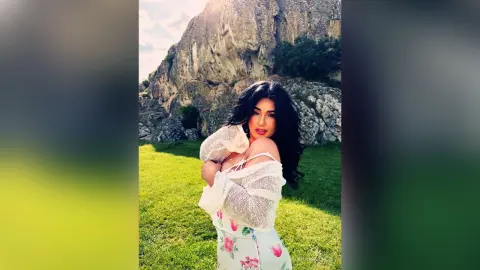 Smiling woman in a white floral dress posing in a sunny green meadow with rocky mountains in the background.