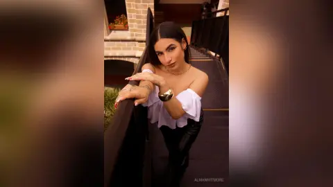 AlmaWhitmore poses outdoors on a balcony wearing a white off-the-shoulder top and black pants, looking up at the camera with long dark hair.
