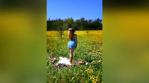 Young woman in a blue summer dress standing in a sunny yellow wildflower field under a clear blue sky.