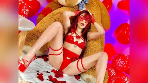 Woman in red lace lingerie relaxing against a giant teddy bear surrounded by Valentine's Day heart balloons.