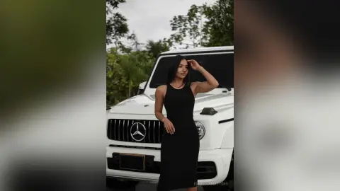 Elegant woman in a sleek black dress standing by a white luxury SUV in a lush outdoor setting.