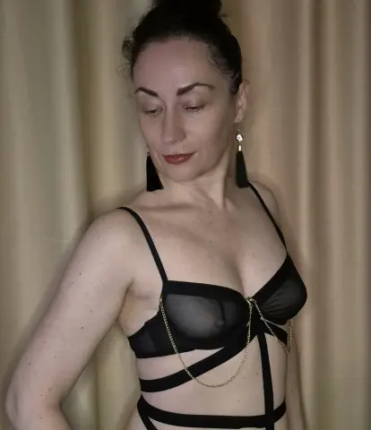 Woman in black sheer lingerie posing against beige curtains in a moody indoor setting.