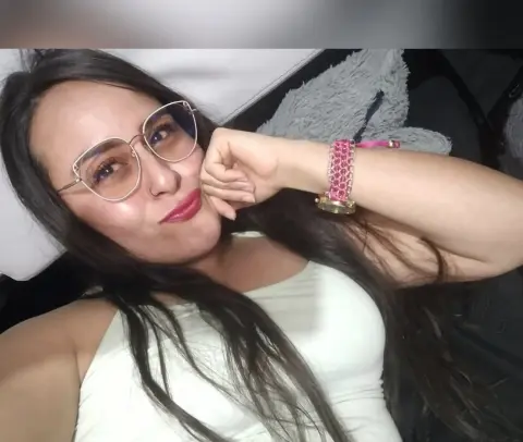 Smiling woman with long dark hair and glasses relaxing in a white tank top at home.