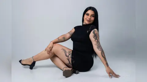 BellaMonrow sits on a white floor wearing a short black dress, showing off her long dark hair and many tattoos on her arms and legs.