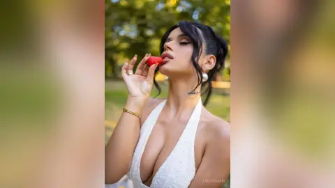 Elegant woman in a white plunging dress eating a strawberry in a sunny outdoor garden setting.