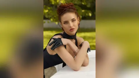 AnjeNoir poses outdoors in a black top and black gloves, looking at the camera with her red hair tied up.