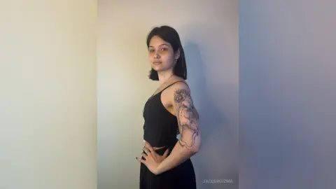 JacquiKozma poses sideways wearing a black tank top that shows off the tattoos on her arm and shoulder.