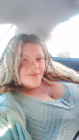 Smiling woman with long blonde dreadlocks wearing a light blue knit top inside a bright car.