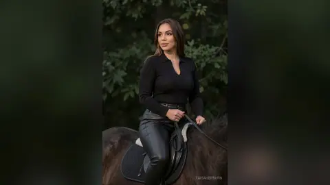 Elegant woman in black equestrian outfit riding a horse in a lush green forest setting.