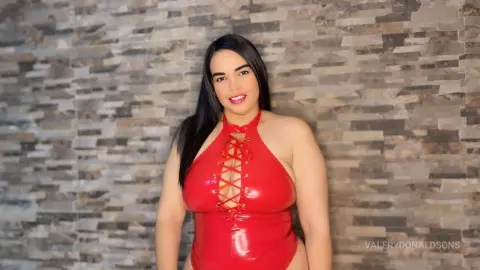 ValeryDonaldsons wears a red shiny lace-up bodysuit while posing against a stone wall background.