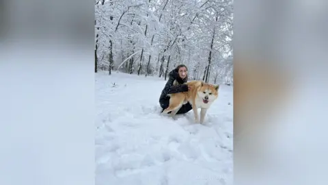 Smiling woman in a black winter coat playing with a fluffy Shiba Inu dog in a snowy winter forest.