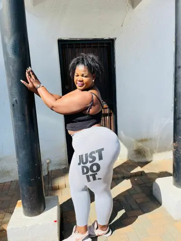 Confident woman in black tank top and grey Just Do It leggings posing outdoors by a pillar in bright sunlight.