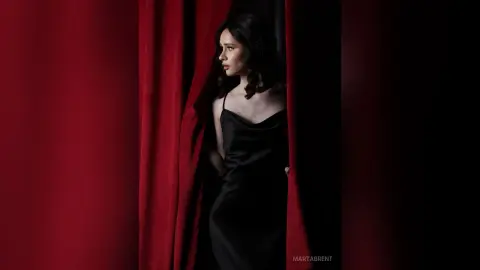 Elegant woman in a black satin slip dress peeking through red velvet curtains in a moody, cinematic setting.