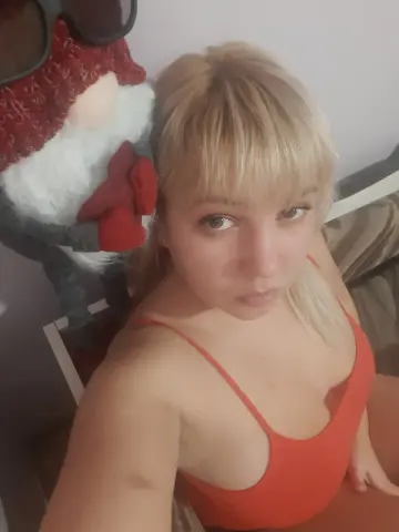 Blonde woman in a red tank top posing indoors next to a large whimsical gnome plushie.
