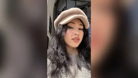 Young woman with long dark hair wearing a beige beret and patterned sweater in soft natural lighting.