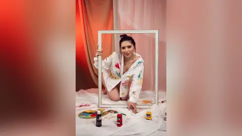 Smiling woman in a white shirt sitting on a bed surrounded by colorful paint bottles in a bright studio.