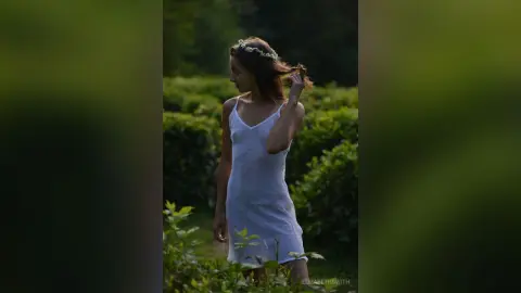 Dreamy woman in a white summer dress and flower crown standing in a lush green garden.
