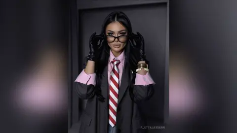 Stylish woman in a pink shirt, striped tie, and black blazer wearing glasses and leather gloves in a studio setting.