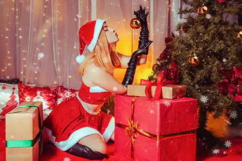 Blonde woman in red Santa outfit sitting by Christmas tree and gifts in a festive holiday setting.