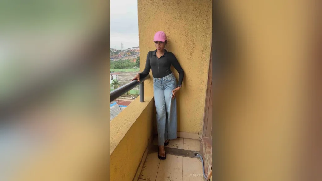 AlbertNina — AlbertNina stands on a balcony wearing a black long-sleeve shirt, light blue jeans, and a pink baseball cap.
