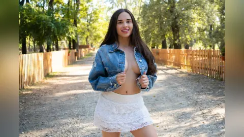 Smiling brunette woman in a denim jacket and white lace skirt walking on a sunny forest path.