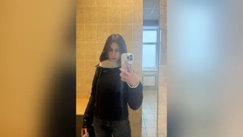 Young woman in a black hoodie and dark jeans taking a mirror selfie in a modern tiled bathroom.
