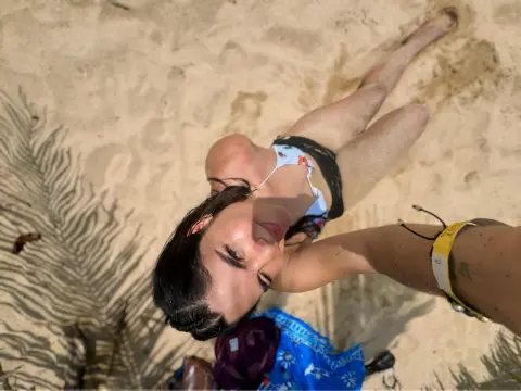 RenataDomick lies on the sand wearing a white floral bikini top, looking up at the camera with dark hair swept back.