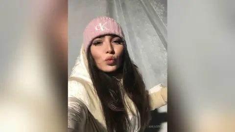 Young woman in a pink Calvin Klein beanie and white winter jacket making a playful pouty face indoors.
