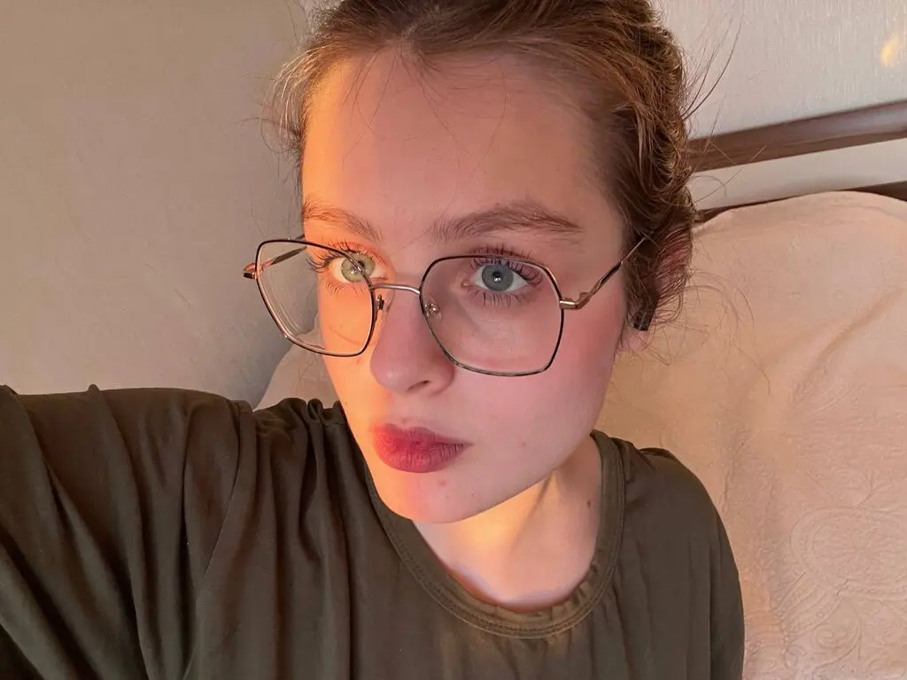 Young woman with glasses wearing an olive green shirt in a cozy, warm-lit bedroom setting.