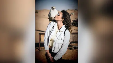 AnaMathers tilts her head back to kiss a light-colored bird perched on her shoulder while wearing a white denim jacket.